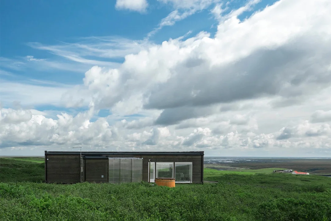 A minimalist black cabin sits isolated in a vast, grassy Icelandic field under a moody sky.