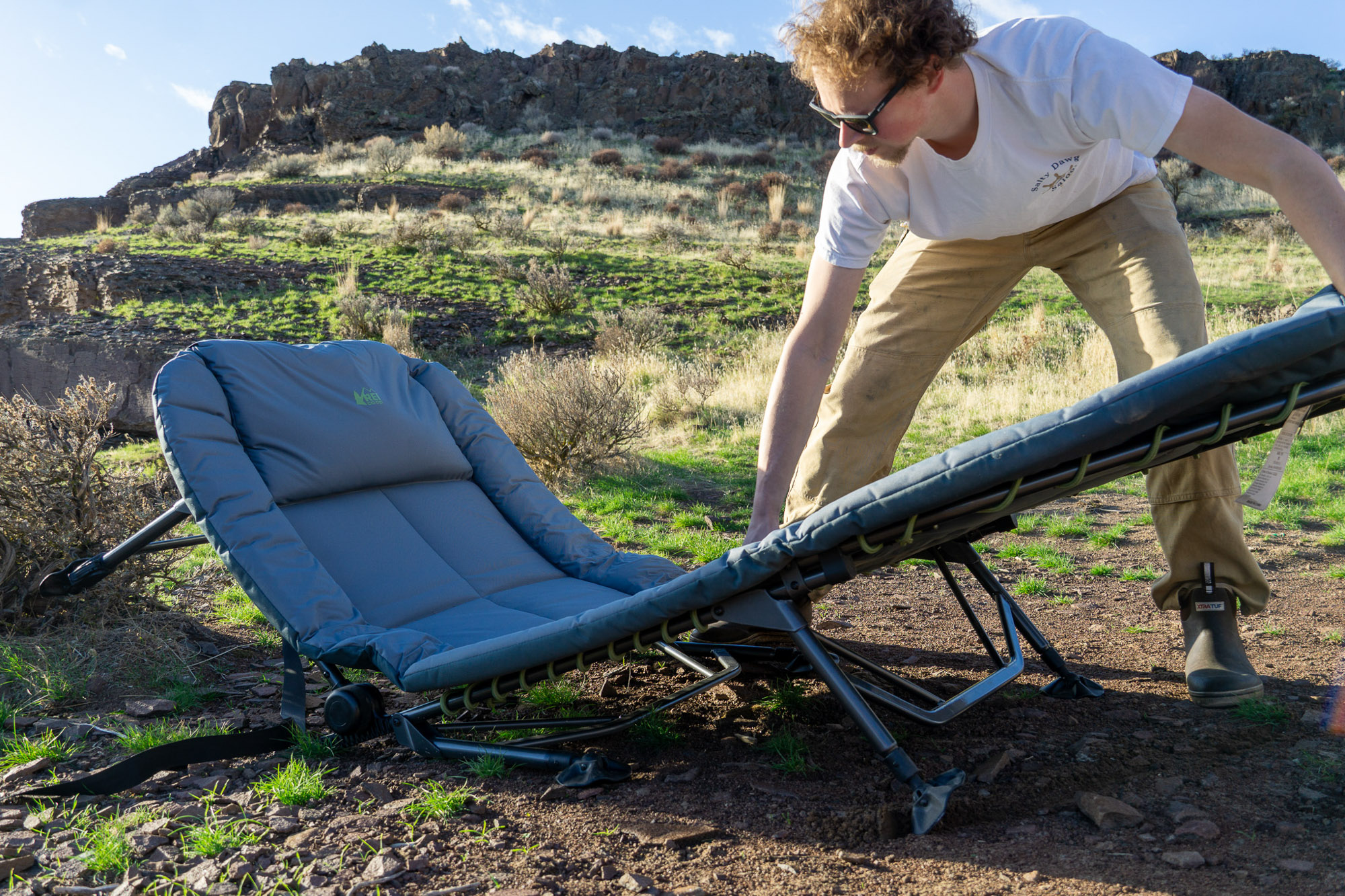 A person unfolding and setting up the REI Wonderland Comfort Cot in a grassy area at an outdoor campsite.