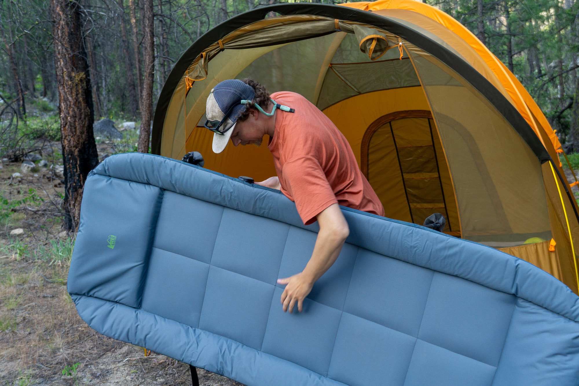 The author carrying the folded REI Wonderland Comfort Cot toward a tent at a campsite.
