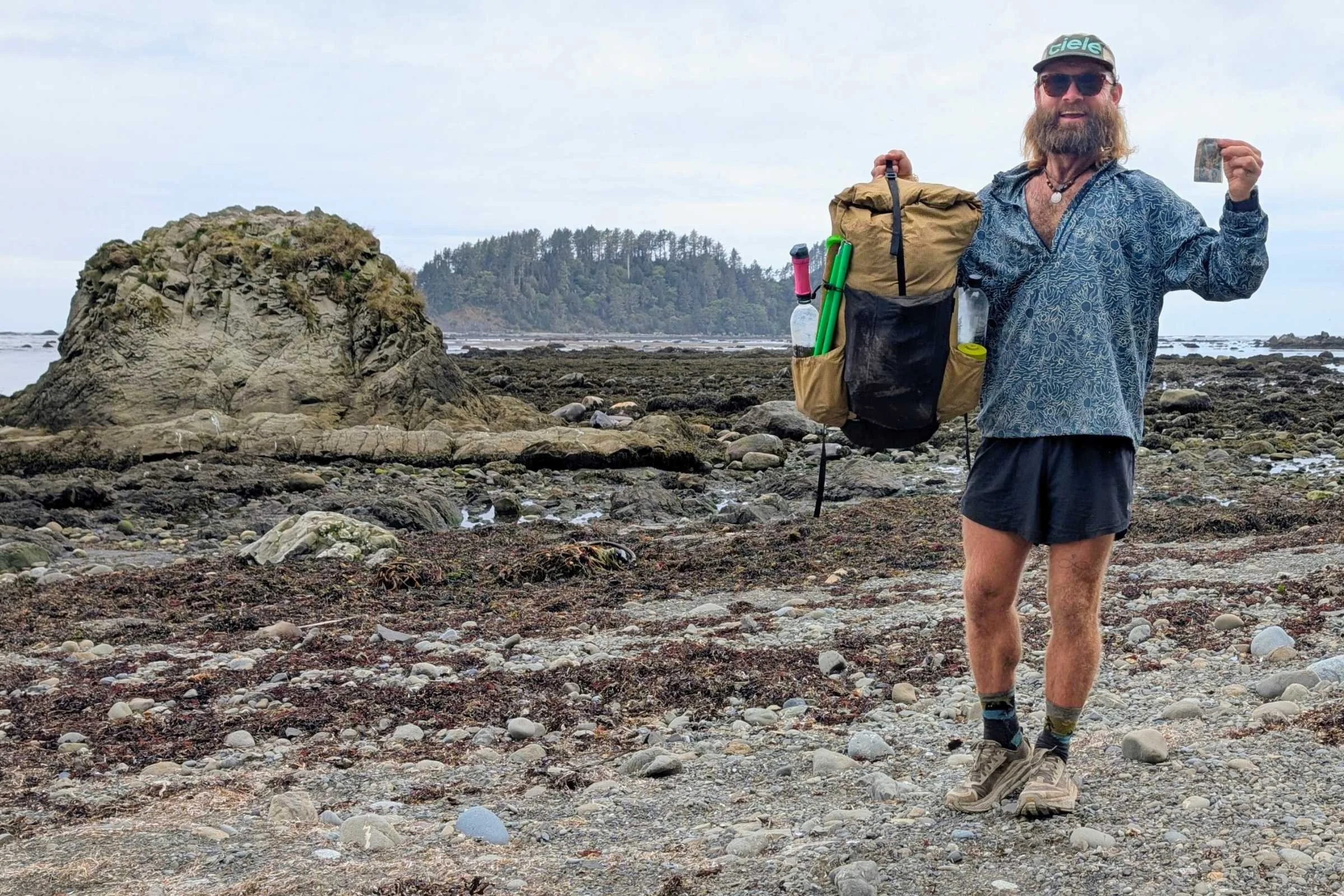 A backpacker walking along a rugged coastline with a bear canister strapped to the top of their pack.