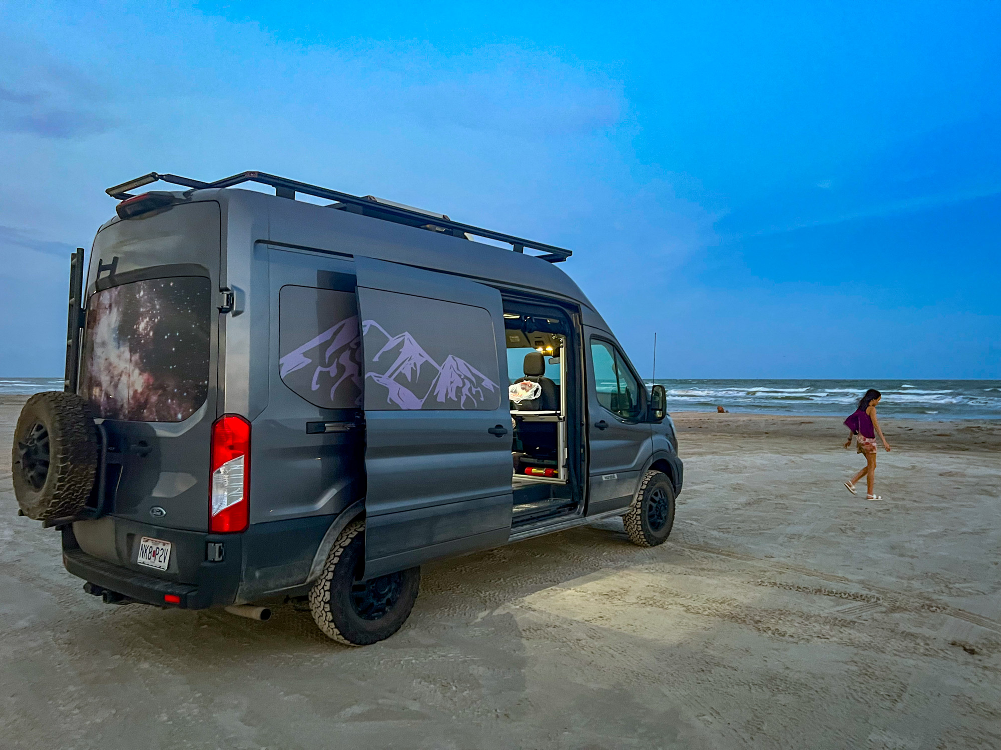 A lifted white campervan parked on a sandy beach during a sunset.