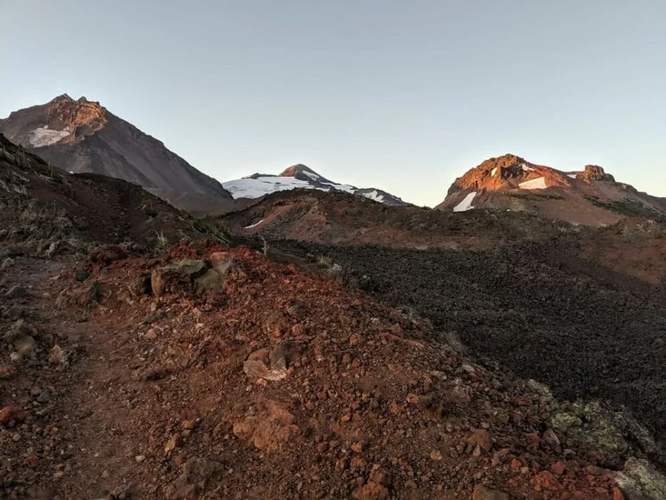 A hiker walking across a rugged, dark lava field with volcanic peaks in the background.